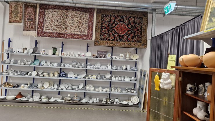 In einem Raum sind Regale mit verschiedenen Keramikwaren und Geschirr zu sehen. An der Wand hängen mehrere Teppiche, darunter ein roter Orientteppich. Rechts steht ein Holzschrank mit weiteren Keramiken und einer gelben Figur.