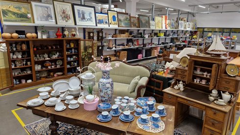 Ein Raum mit antiken Möbeln und Geschirr. In der Mitte steht ein Tisch, der mit verschiedenen Tellern, Tassen und einer dekorativen Vase bedeckt ist. Im Hintergrund sind Regale mit Bildern und weiteren Ausstellungsstücken zu sehen.