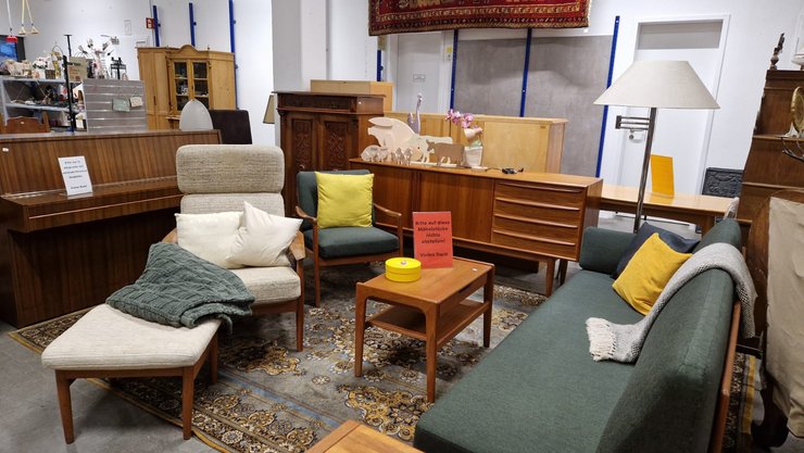 Ein gemütliches Wohnzimmer-Arrangement mit einem grünen Sofa, einem Sessel und einem Liegesessel. Verschiedene Holzmöbelstücke, darunter ein Beistelltisch und ein Regal, sind im Hintergrund zu sehen. Einige Kissen und eine Decke liegen auf den Möbeln.