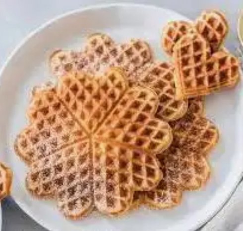 Ein Teller mit mehreren Waffeln in verschiedenen Formen, darunter eine große blütenförmige Waffel und eine kleinere herzförmige. Die Waffeln sind goldbraun und mit Puderzucker bestäubt.