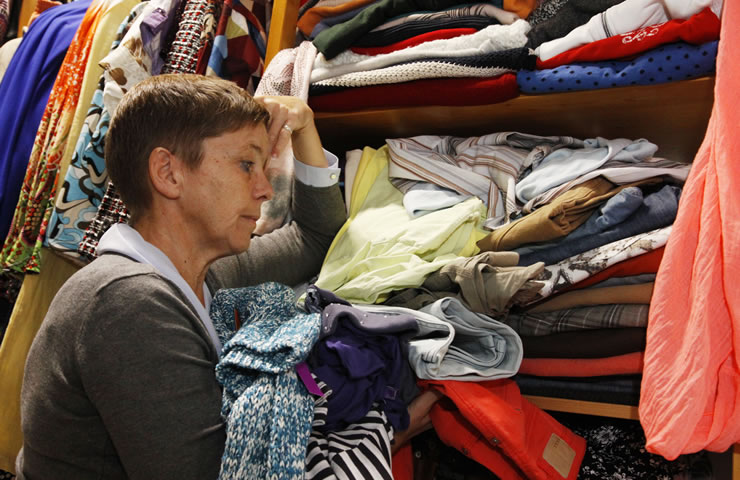 Eine Frau steht an einem Kleiderschrank und schaut nachdenklich auf einen Haufen Kleidung, den sie in den Händen hält. Der Schrank ist voll mit verschiedenen Kleidungsstücken. 
